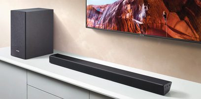 Samsungs sorte, rektangulære, slanke og langstrakte soundbar HW-R530, placeret på et hvidt møbel under en stor vægmonteret skærm, der viser et orange, klippefyldt landskab