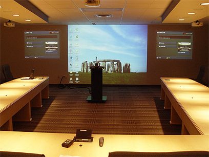 Konferenční místnost s projekčním plátnem zobrazujícím pracovní plochu Windows, dvěma světlými dřevěnými stoly a černým pultem
