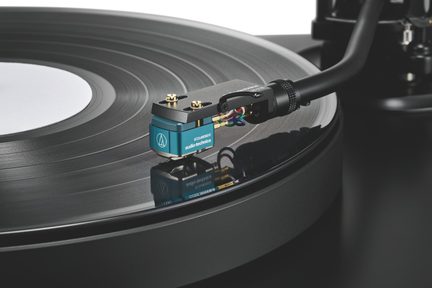 Modrá kovová phono přenoska na černém raménku, jehla na černém vinylu s viditelnými drážkami, šikmý pohled