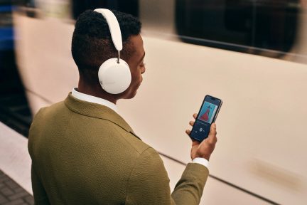 Balti krāsas audio aurēši, melni krāsas smartfons, zaļas krāsas veste, metrosta stacijas platforma, kāda attālumā redzama