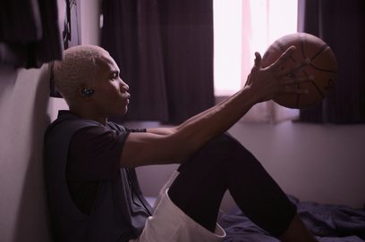 Ung sort dreng sidder med en brun basketbold, iført sort t-shirt, i et rum med lyserøde gardiner