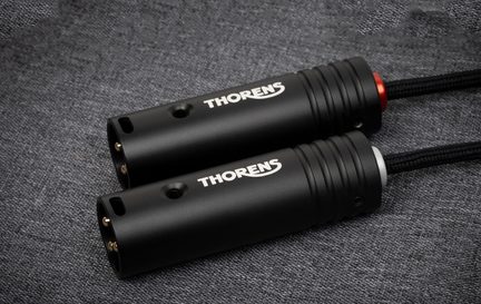 Dva válcové kovové audio konektory v matné černé barvě zobrazené v mírně skloněném pohledu, s bílým logem „THORENS“, třemi pozlacenými kolíky a zadním kroužkem v červené nebo stříbrné barvě, položené na texturované šedé látce.