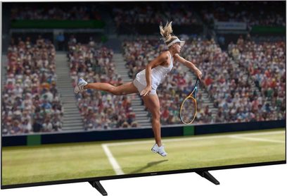 Panasonic TX-65JX940E-fladskærm, der viser en kvindelig tennisspiller i hvid dragt, der springer med løftet ketsjer på en græsbane foran en tilskuerskare