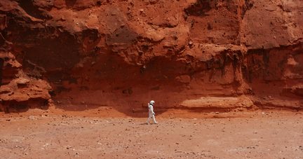 Wide side view of a person in a white spacesuit with a smooth composite helmet, walking on red rocky ground at the foot of a tall orange sandstone cliff