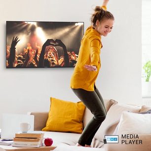 Smilende ung pige i gul sweater, der glade hopper foran et TV, der viser en koncert med publikum, der løfter armene