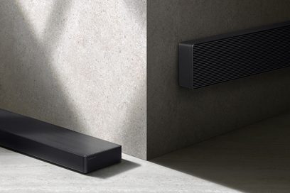 Černý zvukový panel z matného kovu s vodorovným drážkovaným mřížkováním, tenký obdélníkový tvar se zaoblenými rohy, pohled ze tří čtvrtin ukazující jeden modul položený na zemi a druhý upevněný na stěně