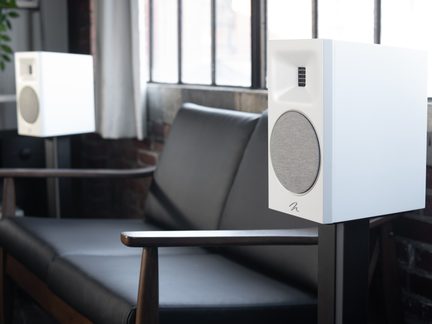 Two white speakers on black stands, a black sofa with wooden armrests, windows with metal frames in the background
