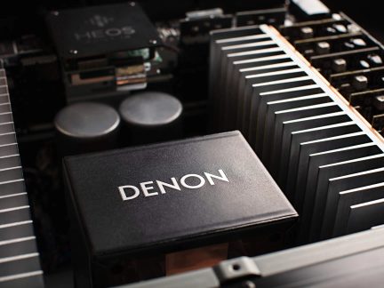 Detailní pohled shora na rámec audio zesilovače, černý kovový kryt s nápisem Denon, doplněný stříbrnými hliníkovými chladicími žebry a dvěma válcovitými šedými kondenzátory