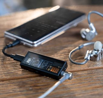 Rektangulært svart kabinett med synlig krets, tilkoblet kabel, sølvfargede ørepropper med hvite tupper, svart smarttelefon på skrå