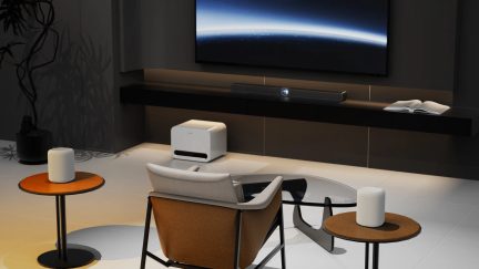Salotto moderno con videoproiettore bianco a ottica corta di fronte a uno schermo a parete, soundbar e poltrona in primo piano