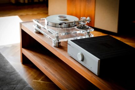 Transparent pladespiller med forkromet tonearm og sølvfarvet hi-fi-forstærker på træmøbel
