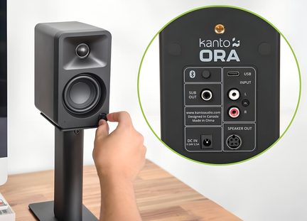 Kompakt sort højttaler i mat plast, set fra trekvart forfra på stativ med en hånd, der drejer på lydstyrkeknappen på fronten, cirkulær overlejring viser detaljer om lyd-, USB- og strømporte på bagpanelet