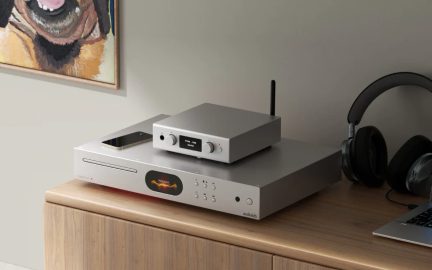 Dva obdĺžnikové hi-fi audio prístroje zo strieborného brúseného kovu, jeden so štrbinou na optický prehrávač, oválnym displejom a tlačidlami, zatiaľ čo menší, položený na ňom, má centrálny displej, dve otočné ovládače a zadnú anténu, pohľad z pravého predného trojštvrťového uhla, umiestnené na drevenom nábytku.