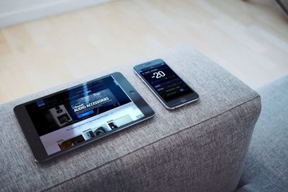 Subwoofer SVS SB-2000 Pro in nero lucido e smartphone che mostra l'app di controllo, appoggiati su un divano grigio
