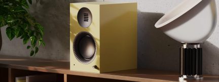 Boxă de raft galbenă cu difuzor negru pe o etajeră din lemn, lângă o lampă albă modernă