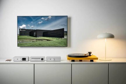 Kollane Pro-Jecti vinüülplaadimängija valgel mööbliesemel koos sobiva hi-fi süsteemi ja põleva laualambiga
