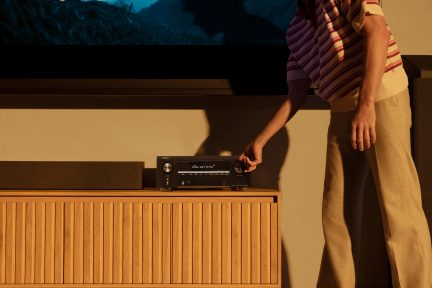 Černý domácí kino zesilovač Denon na dřevěném nábytku, ruka na ovladači hlasitosti, soundbar vlevo