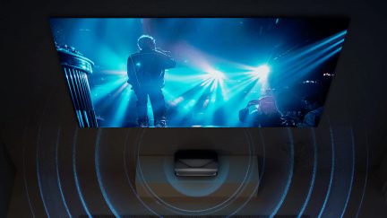 Silhuet af en mand på scenen, oplyst af blå spotlights, foran en skærm der viser et sløret billede af ham selv set bagfra