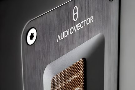 Nærbillede i trekvart vinkel af den grå børstede metalfrontplade på en Audiovector-lydenhed, udstyret med en sort stjerneformet skrue, det ovale logo og den hvide “AUDIOVECTOR”-tekst samt en indfældet rektangulær åbning med gyldent metalgitter