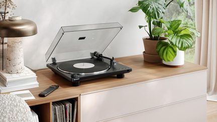 Black vinyl turntable with plastic and metal construction, transparent lid raised, three-quarter view on a wooden furniture piece