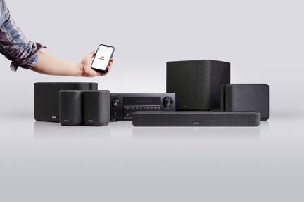 Schwarzes Audio-Set aus Akustikstoff, bestehend aus Soundbar, Subwoofer, zylindrischen Satellitenlautsprechern und Digital-Display-Empfänger, Frontansicht auf grauem Hintergrund, ein Arm links hält ein Smartphone