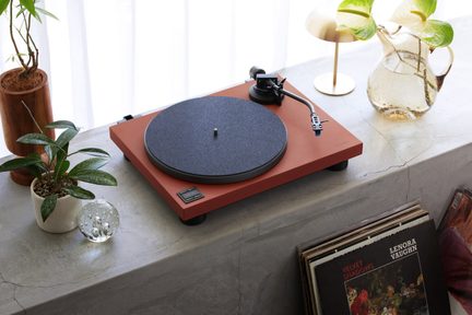 Tocadiscos de vinilo, base de madera probable en rojo ladrillo mate, plato con fieltro gris y brazo en S plateado, vista en tres cuartos