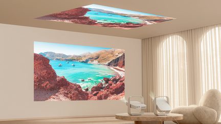 Two white video projectors on a coffee table, projecting a panoramic view of turquoise sea and red cliffs on the wall and ceiling