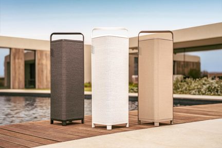 Tres altavoces de columna Bluetooth para exterior en gris, blanco y beige sobre una terraza de madera junto a una piscina