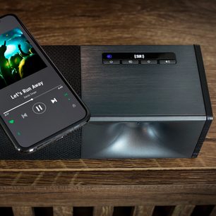 Soundbar Klipsch Cinema 600 neagră cu telecomandă, pe un suport de lemn, afișând opțiuni pe un ecran LED albastru