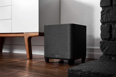 Denon Home Subwoofer svart, kubisk form, svart galler på framsidan, placerad på parkettgolv nära en vägg