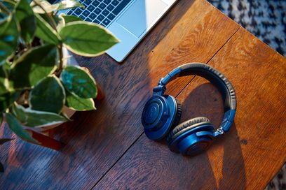 Sluchátka Audio Technica ATH-M50xBT2DS v modro-stříbrné barvě položená na dřevěném stole vedle notebooku a zelené rostliny