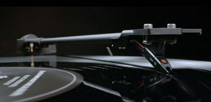Sort tonearm med pickup og nål i kontakt med en sort vinylplade, nærbillede fra en sidevinkel.