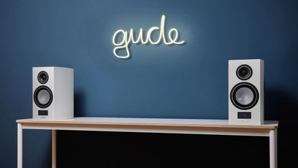Sæt hvide Bluetooth-højttalere på et lyst træskrivebord, blå væg med neonskiltet “gude” i kursiv skrift