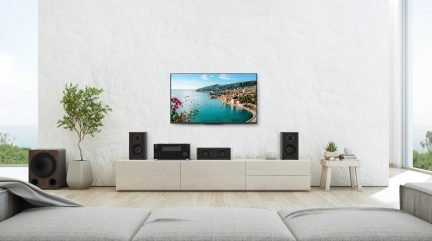 Beyaz dokulu duvarlı salon, kıyı manzarası gösteren duvar televizyonu, açık renkli alçak mobilya, hoparlörler, amplifikatör, saksı bitkisi
