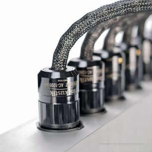 Tæt påbillede, skråt vinkel, af de sorte, blanke plastcylindriske forbindere og de grå-sorte fletkabler, der er placeret på den rektangulære, sølvgrå aluminiumshus med børstet finish på Inakustik AC-3200 Filter Digital SCHUKO-stikdåsen.