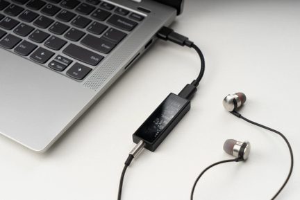 Sølvfarvet tastatur på en bærbar computer, sort lydadapter tilsluttet via kabel, kablede metalhovedtelefoner med grå ørepropper