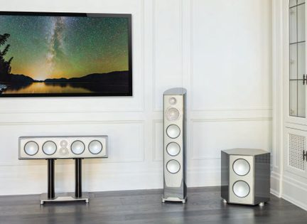 Sæt med tre højttalere i grålakeret metal og hvid, set forfra, bestående af en søjle med fire højttalere, en horisontal centerhøjttaler med fire højttalere monteret på dobbeltfod og en kubeformet subwoofer med to højttalere, reflekterende overflader på mørkt parketgulv foran hvid væg med stuk.