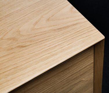 Nahaufnahme der oberen Ecke eines Möbelstücks aus hellem Holz, mit ausgeprägter Maserung, seidenmatter Oberfläche und abgeschrägter Kante