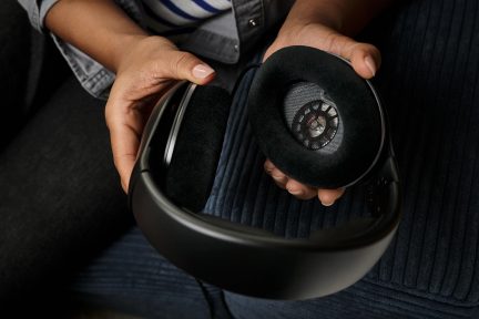 Twee handen houden een zwarte koptelefoon vast met een verwijderd oorkussen, waardoor een interne ronde luidspreker zichtbaar wordt.
