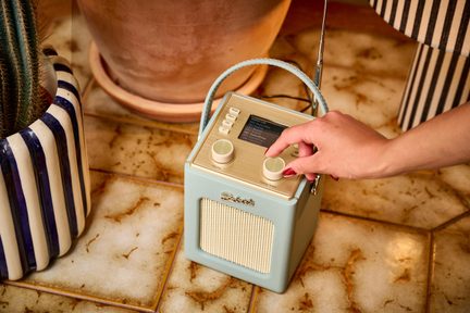 Pastellblaues Radio aus Kunstleder, Ansicht von oben, Display, zwei Knöpfe, cremefarbene Stoffabdeckung, Griff und Antenne, Hand auf einem Knopf