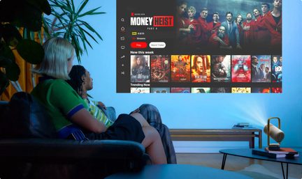 Bílý projektor s plastovým pláštěm a zlatým madlem na stole, projekce rozhraní streamovací služby na zeď, lidé na pohovce