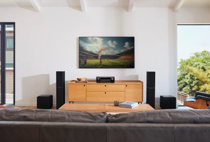 Fladskærms-tv på lyst trækabinet med to sorte højttalere, der viser en fodboldstadion med orange røg på banen