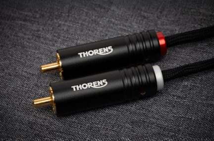 Primo piano laterale di due connettori RCA cilindrici in metallo nero opaco con contatti dorati, anelli rosso e bianco, logo Thorens stampato in bianco e cavo intrecciato nero