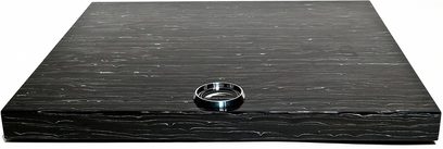 Rektangulær træplade i mørkt rillemønstret træ med en indlejret skinnende metalring i midten, set oppefra