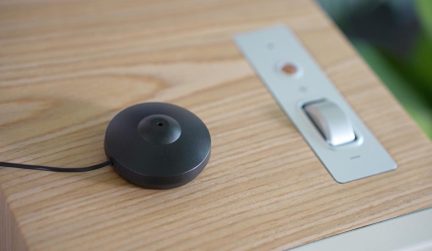 Runder schwarzer kabelgebundener Sensor auf einer hellen Holzplatte, neben einem silbernen Unterputzschalter