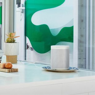 Trådløs, tilsluttet Sonos One (Gen 2) højttaler i mat hvid foran et grønt abstrakt vægmaleri på en hvid hylde