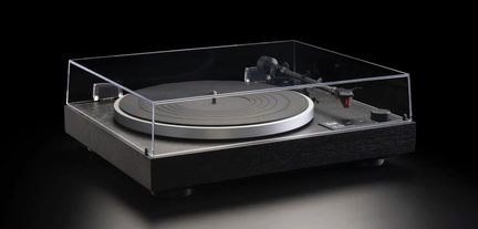 Toca-discos vinil preto e prata, gabinete retangular em madeira escura texturizada, prato metálico circular, tampa acrílica transparente fechada, braço de leitura preto com cápsula vermelha, vista em três quartos pela frente levemente de cima