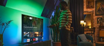 Black flat-screen TV with a slim glass and plastic frame, left oblique view, placed on a low cabinet, green and blue LED backlighting diffused on the wall