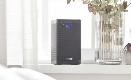 Kompakt grå Canton WiFi-højttaler med blåt LED-display placeret på en moderne vindueskarm ved siden af en glasvase
