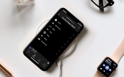 Sort smartphone i glas og metal set oppefra, placeret diagonalt på en hvid trådløs oplader med kabel, ledsaget til højre af et rosaguld smartwatch med beige rem og omgivet af tilbehør på hvid baggrund